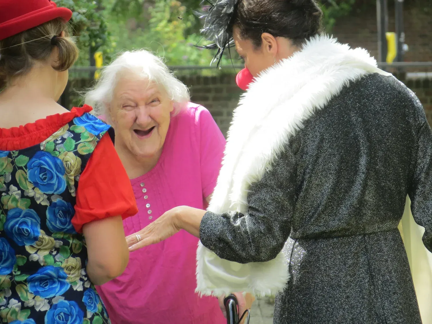 Smiling woman in pink greeting two costumed people outdoors, one in a red cap and one in a black-and-white outfit