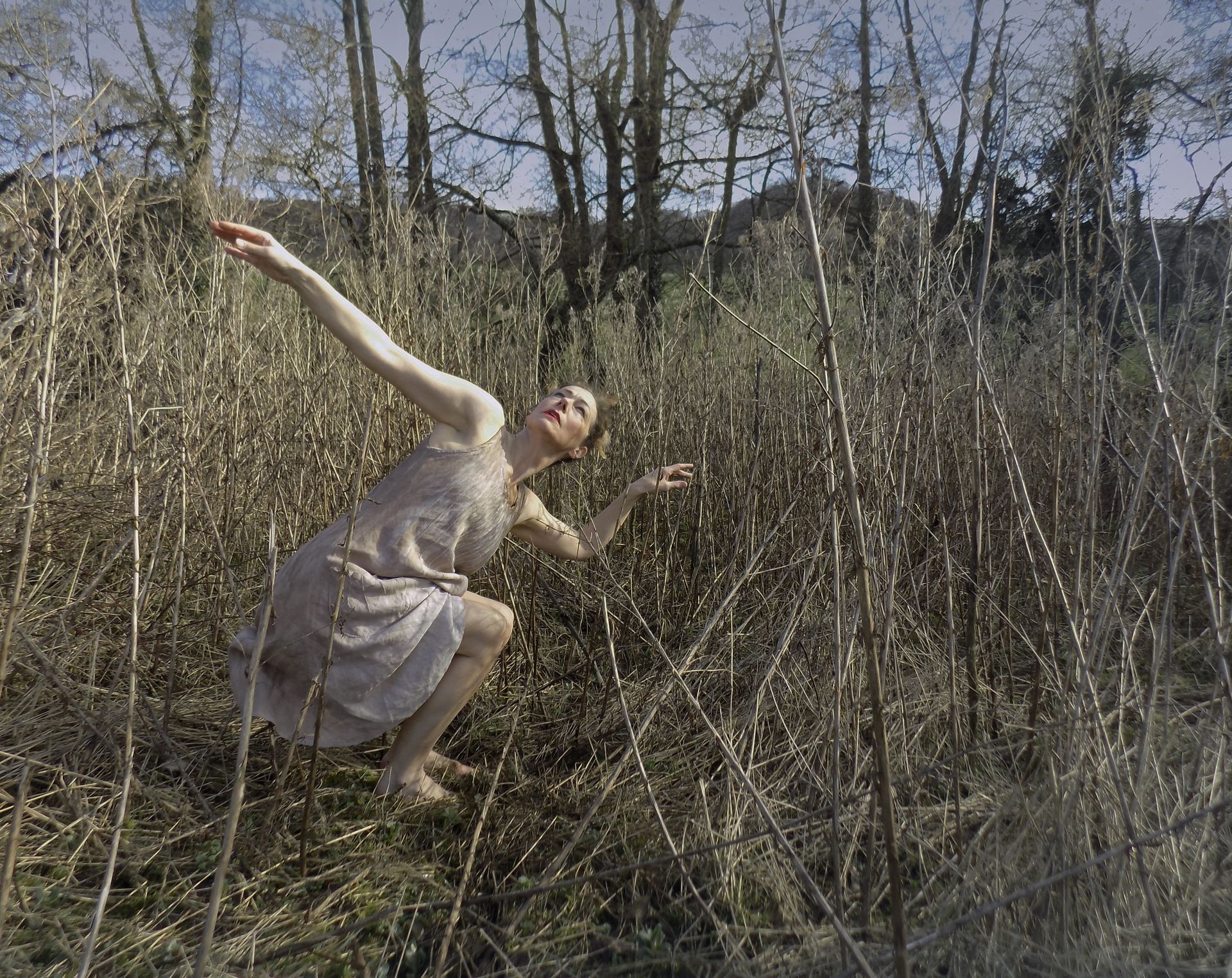 Person in a light dress posing playfully in tall dry grass under bare trees outdoors.