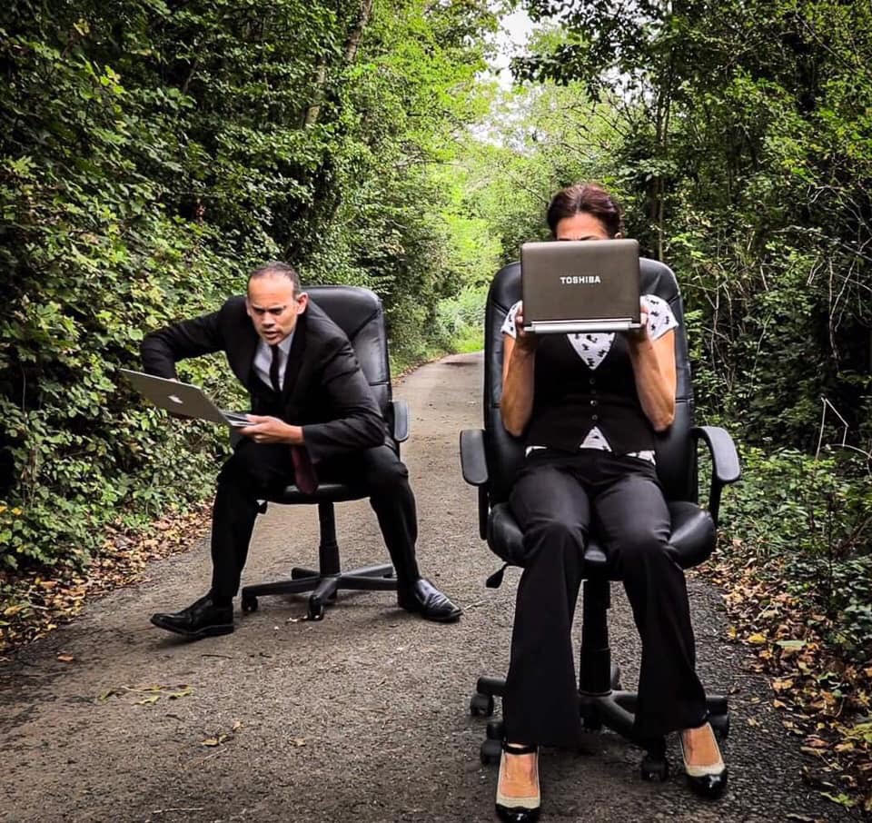 Two people in suits sit outdoors on a narrow path, one using a laptop on their lap.
