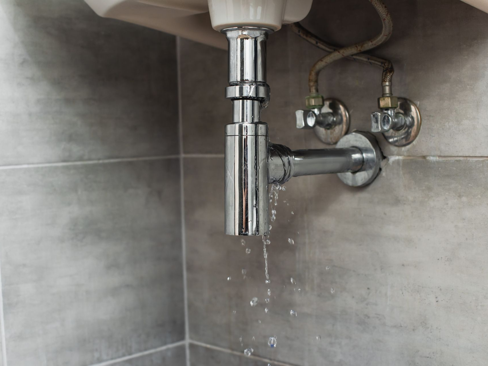 Leaking chrome sink drain pipe under a bathroom sink, with water dripping onto gray tile.
