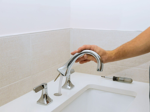 Hand turning on a chrome bathroom faucet over a white sink in a tiled corner.