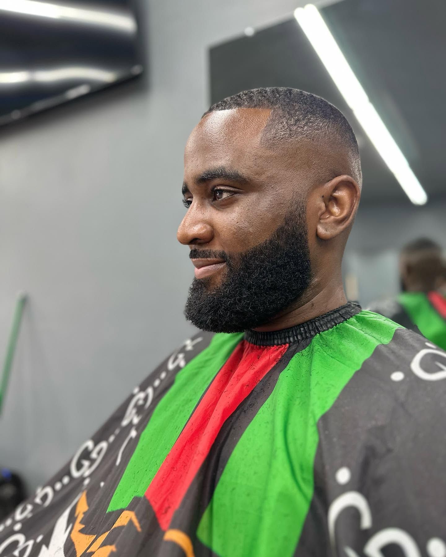 Man seated in a barber chair wearing a black, green, and red cape, with a fresh fade haircut and beard
