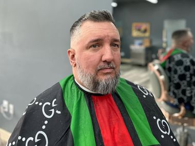 Man seated in a barber chair wearing a black-and-green barber cape, looking off to the side in a salon.