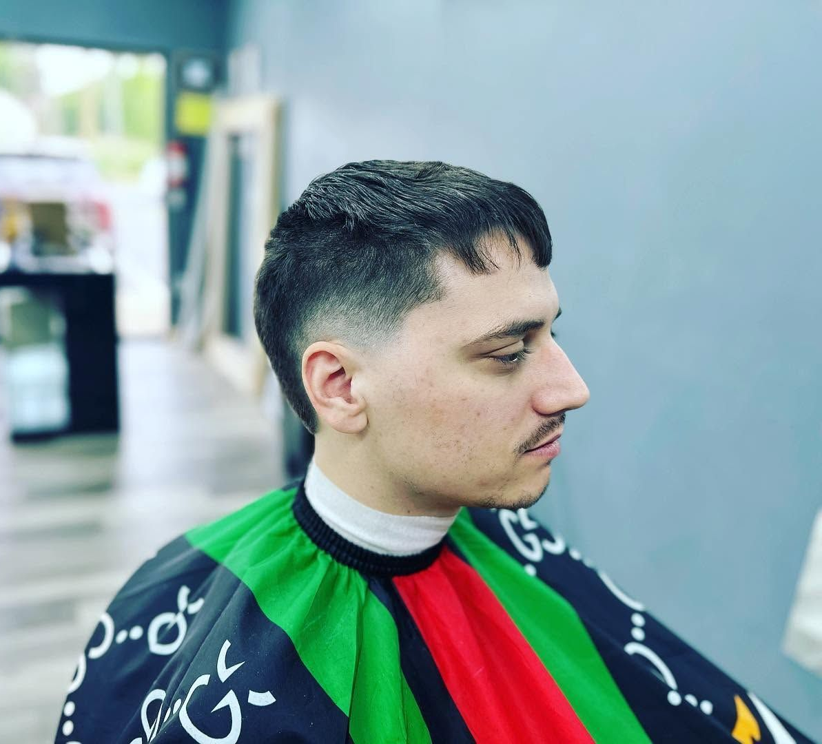 Side-profile portrait of a man in a black, green, and red barber cape, looking ahead in a salon.
