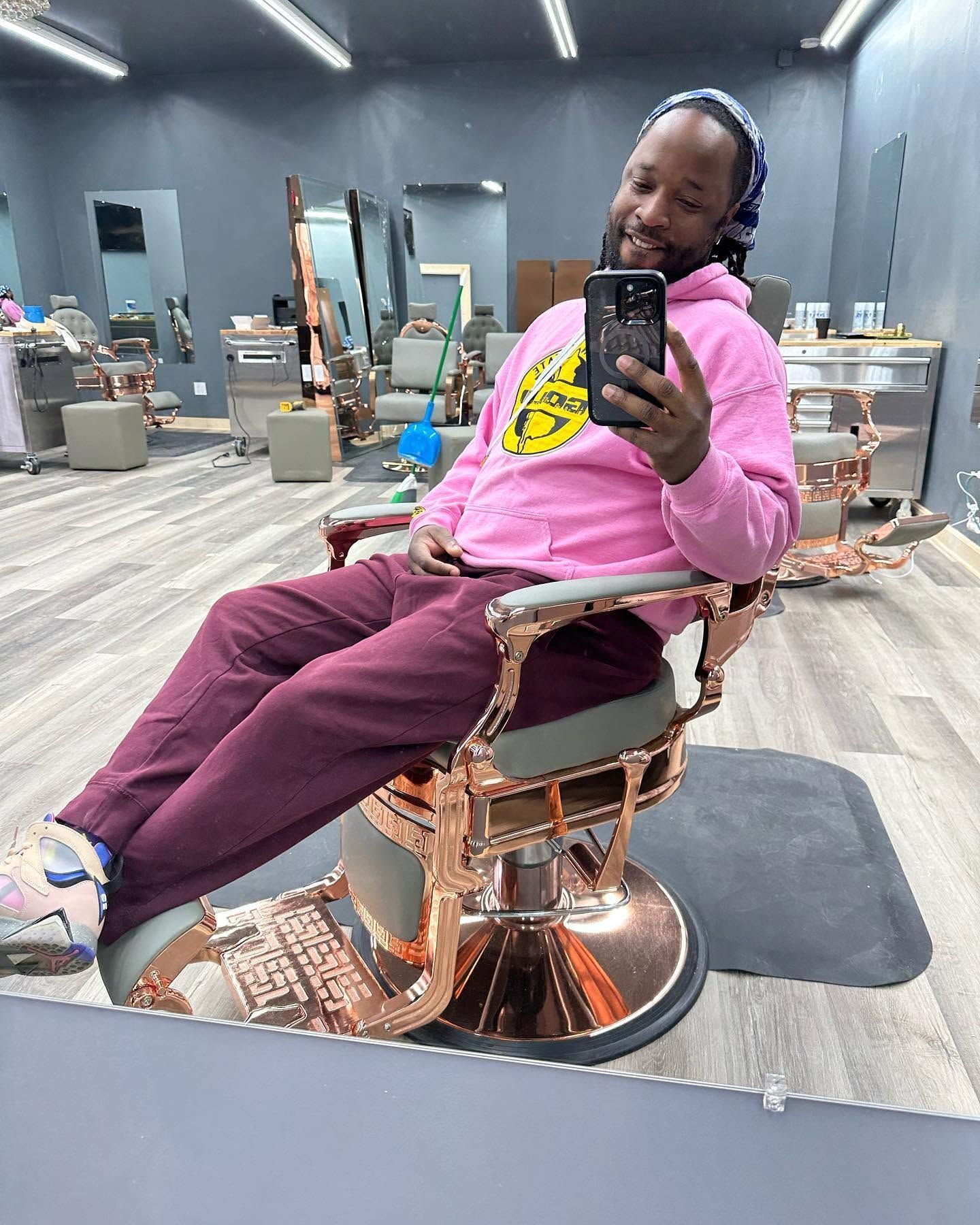 Person smiling in a pink hoodie, taking a selfie in a barber chair inside a bright barbershop.