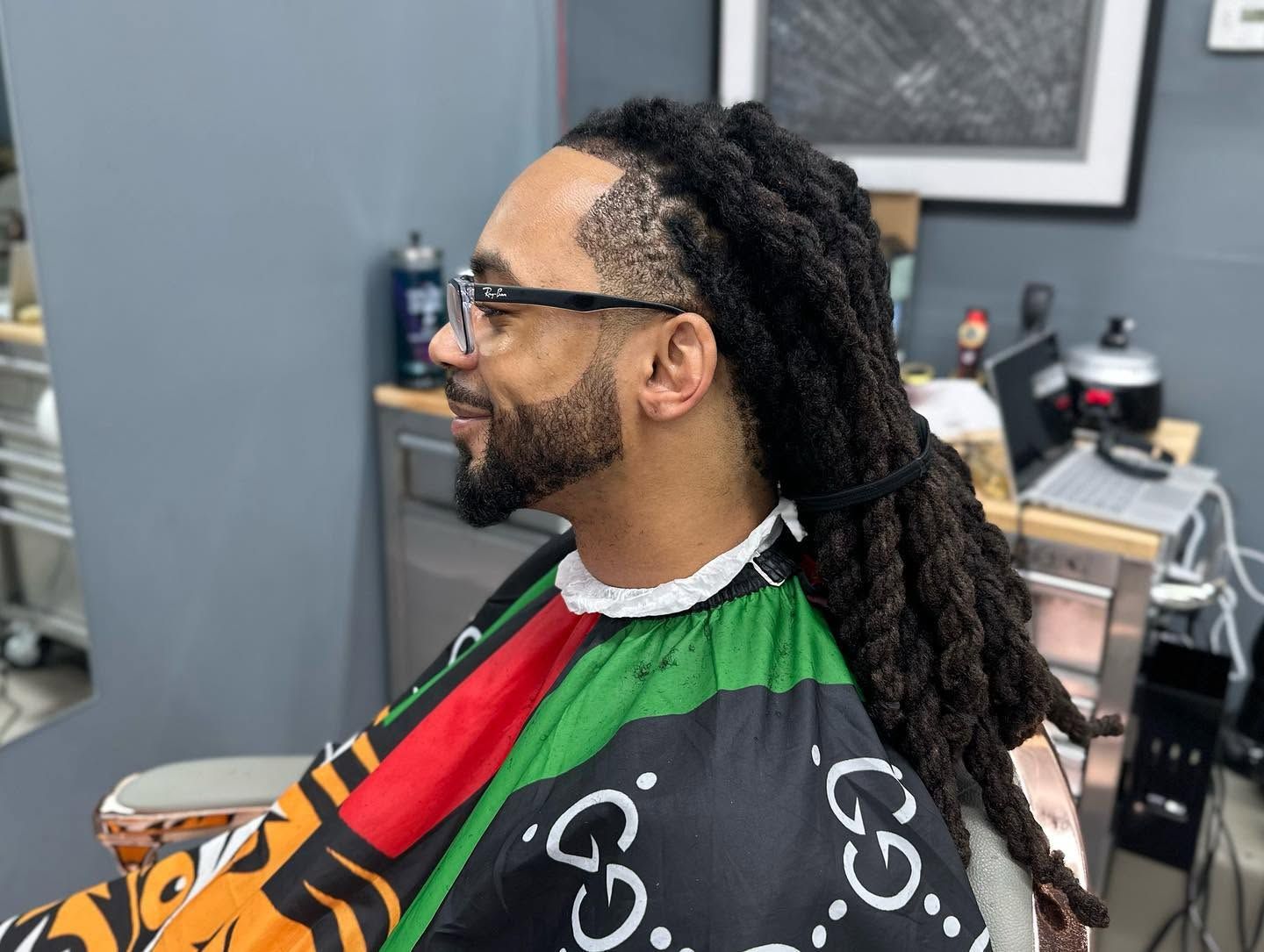 Person with long dreadlocks and glasses seated in a barber chair, wearing a green, red, and black cape.