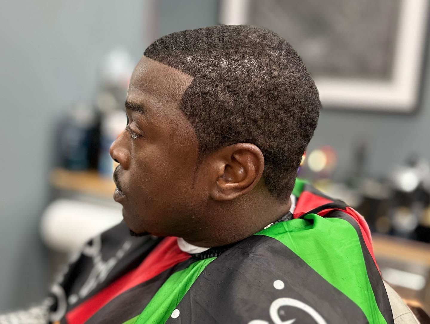 Man seated in a barber chair wearing a black cape with red and green trim, viewed in profile.