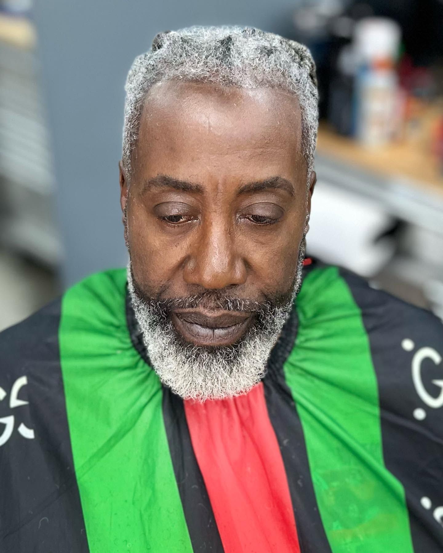 Man with gray beard in a green-and-black barber cape, seated indoors, looking down.