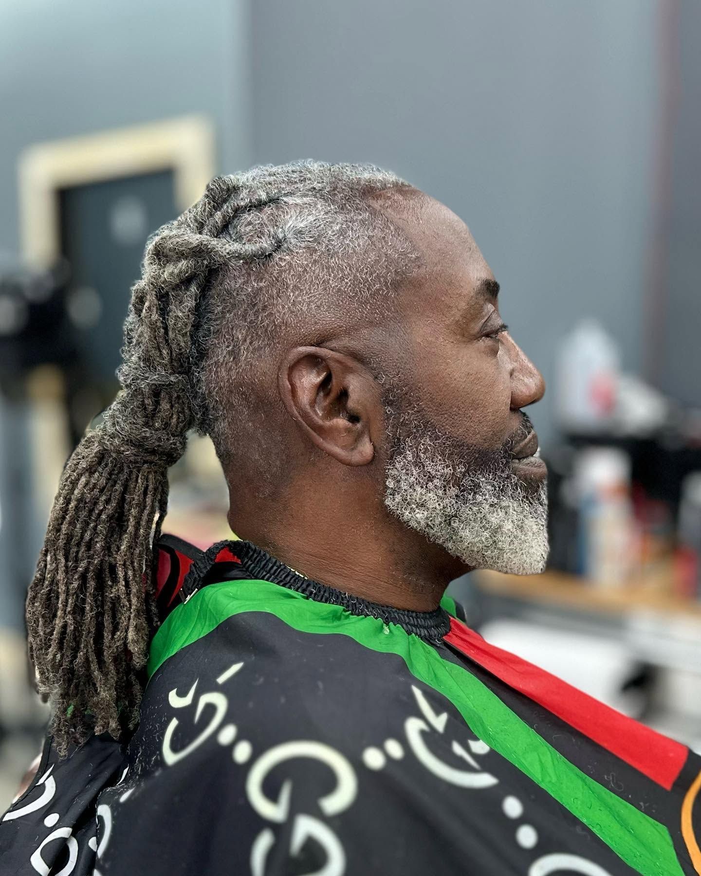 Side profile of a man with gray dreadlocks and beard, wearing a black and green patterned cape in a salon.