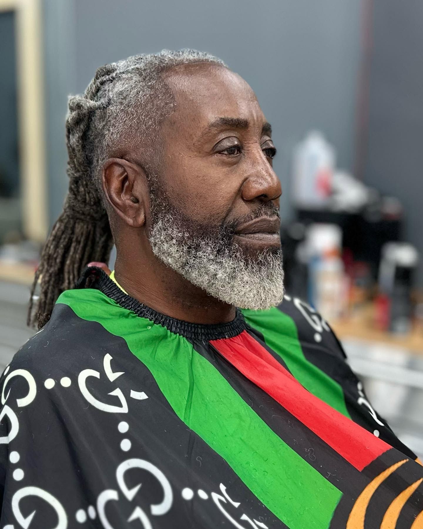 Man seated in a barber chair wearing a black cape with green and red stripes, shown in profile.