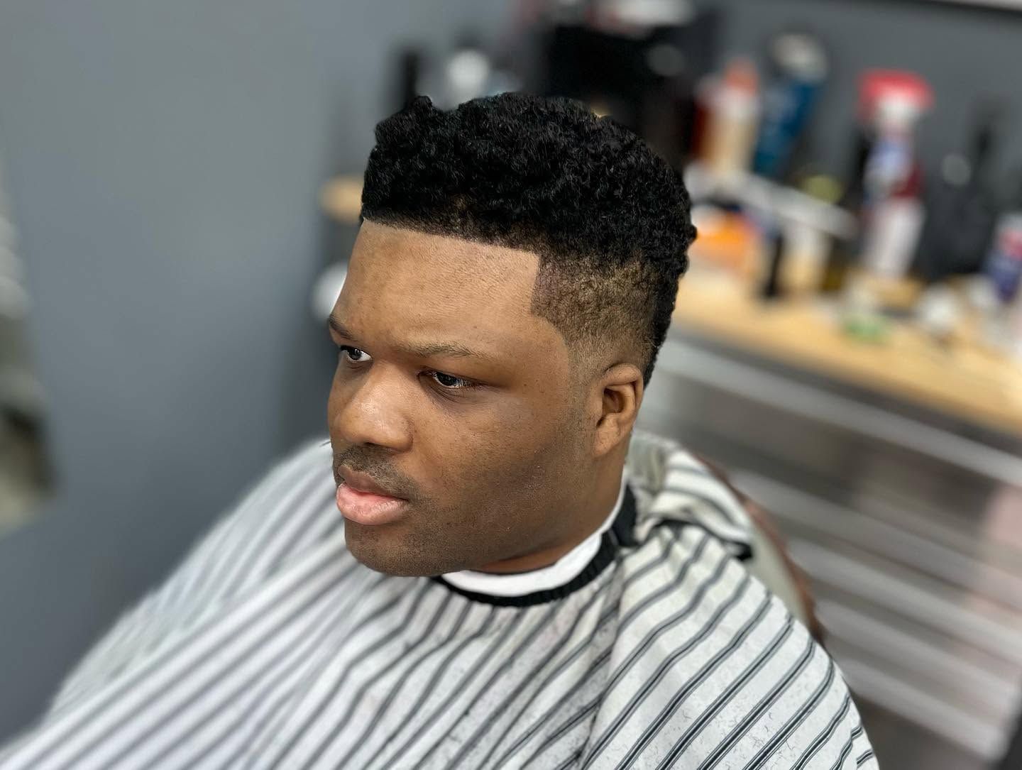 Man in striped barber cape with a fresh fade haircut, seated in a barbershop chair.