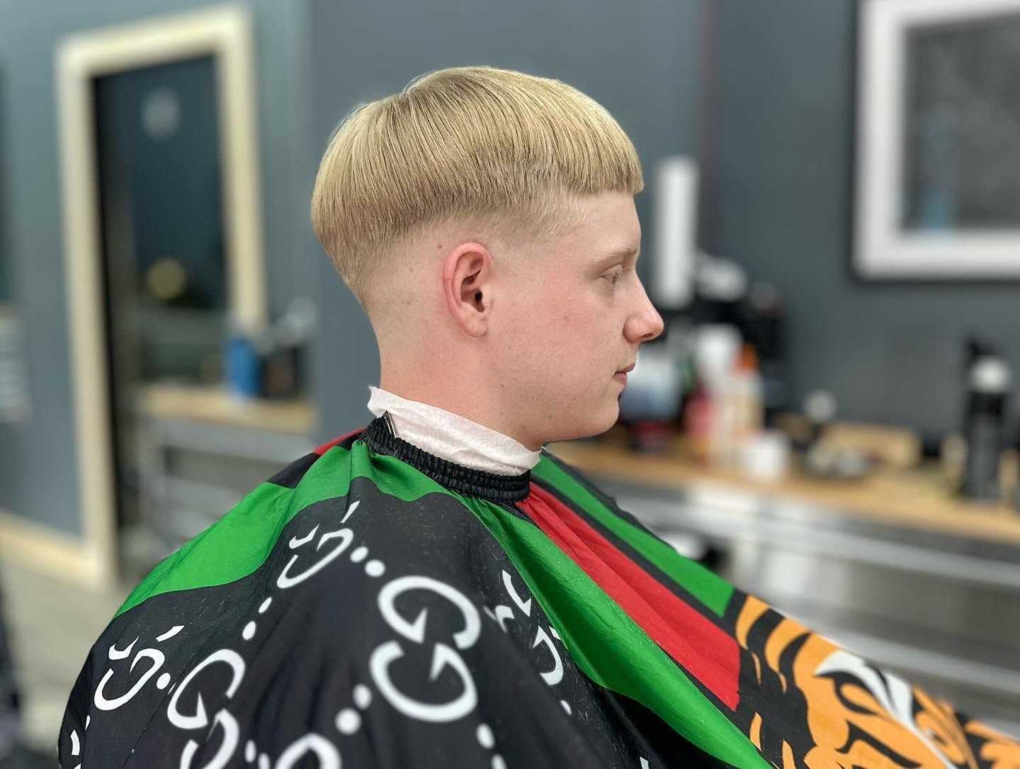 Person getting a haircut in a barbershop, wearing a black cape with green, red, and orange patterns.