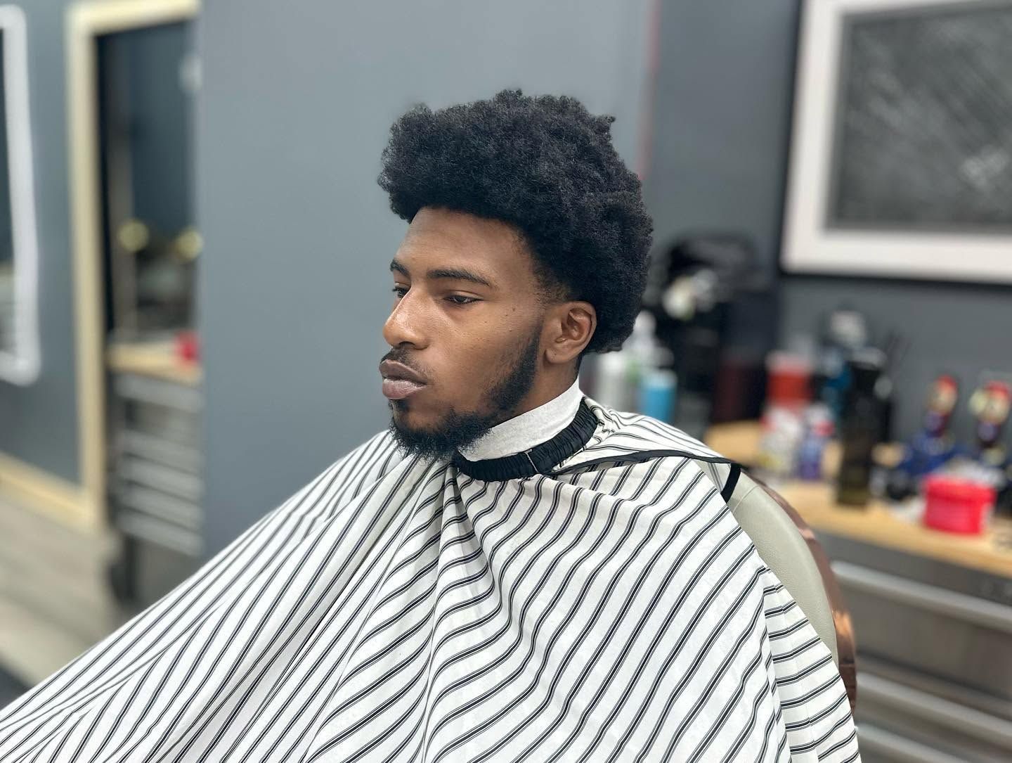 Man in a barber chair wearing a striped cape, with a high curly hairstyle, in a salon setting.