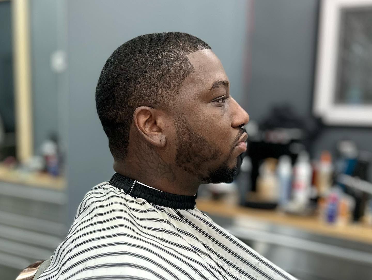 Side-profile portrait in a barber chair, wearing a striped cape inside a barbershop.