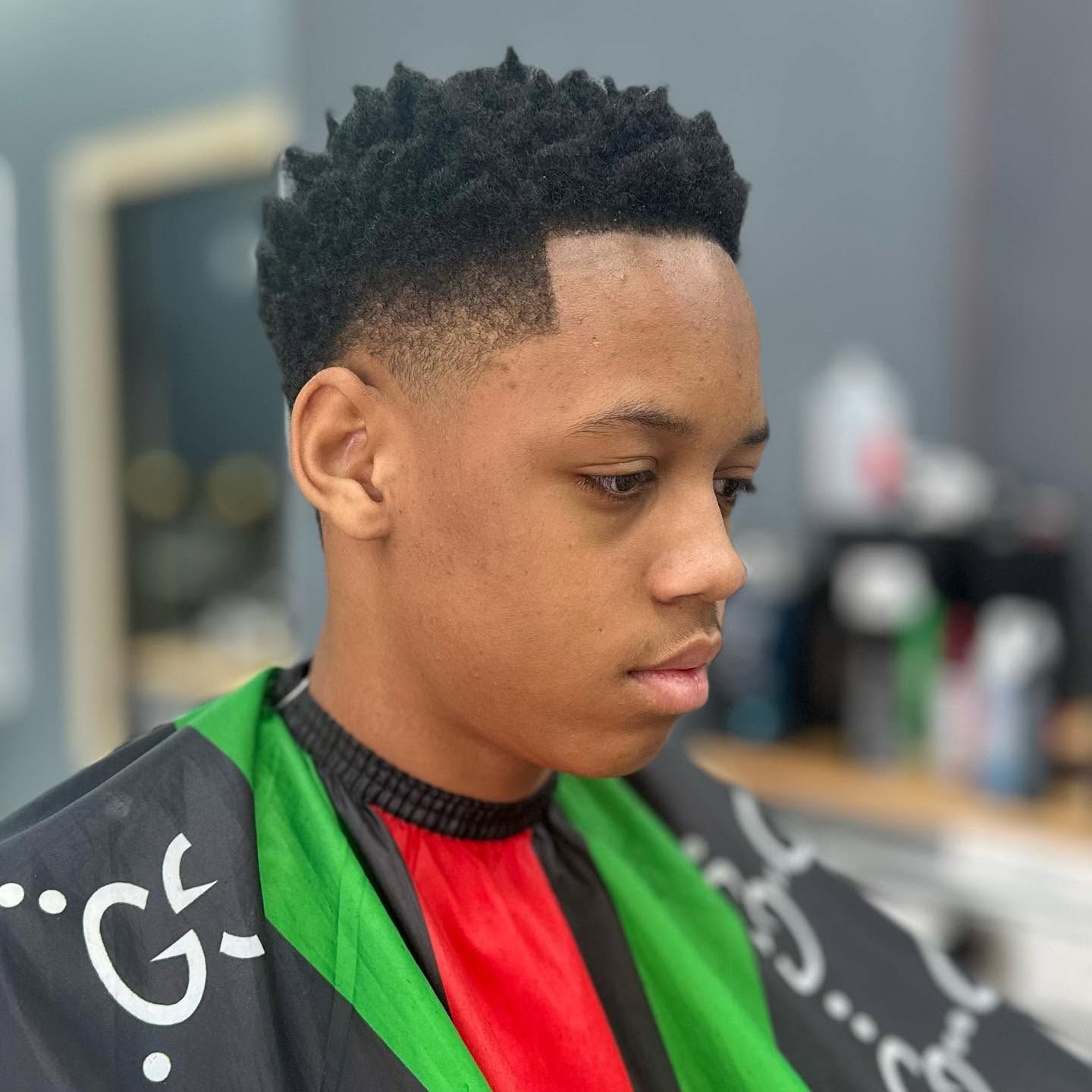 Person with short curly hair sitting in a barber chair, wearing a red shirt and black barber cape with green trim.