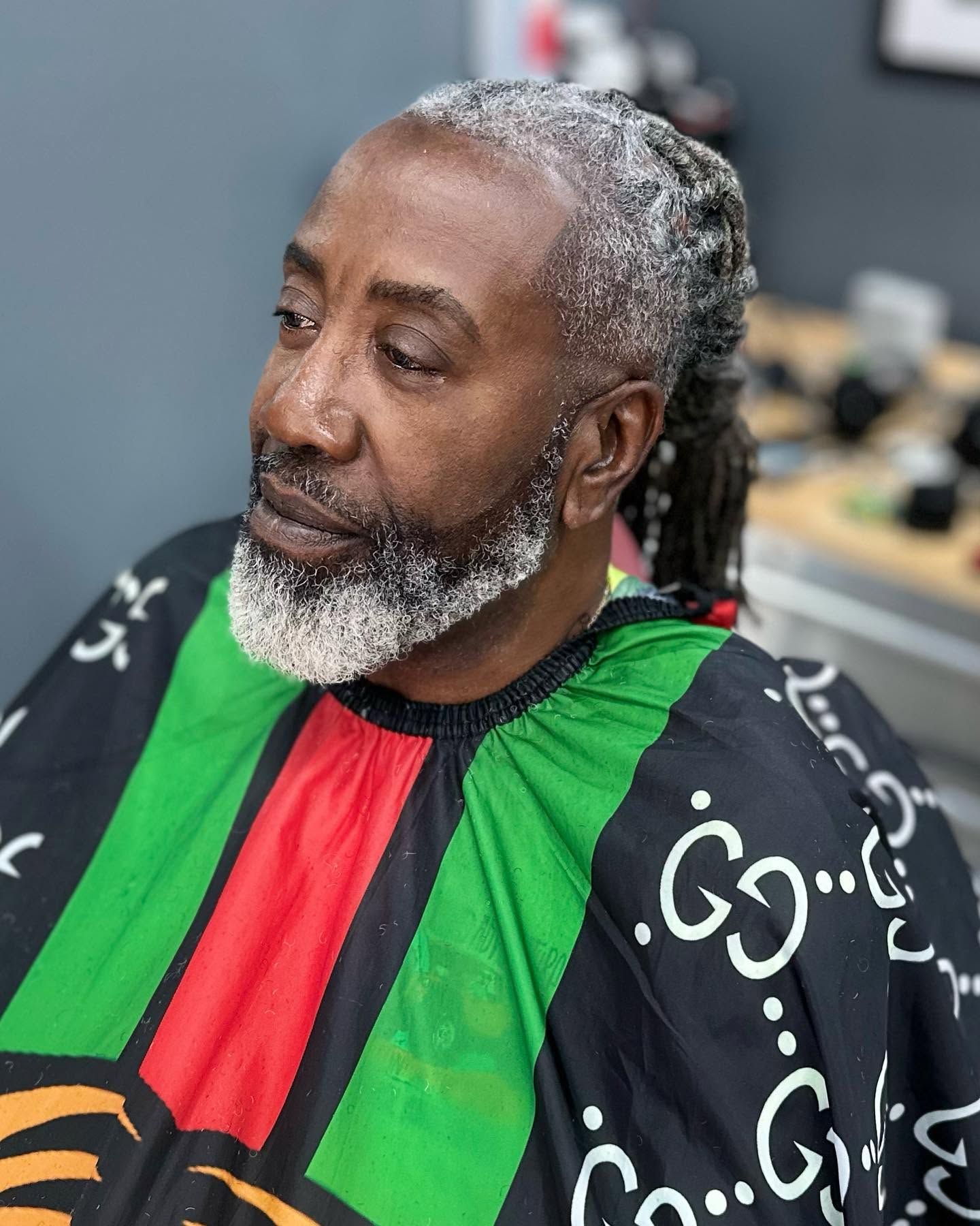 Man seated in a barber chair wearing a green, red, and black cape, with a gray beard and short gray hair.