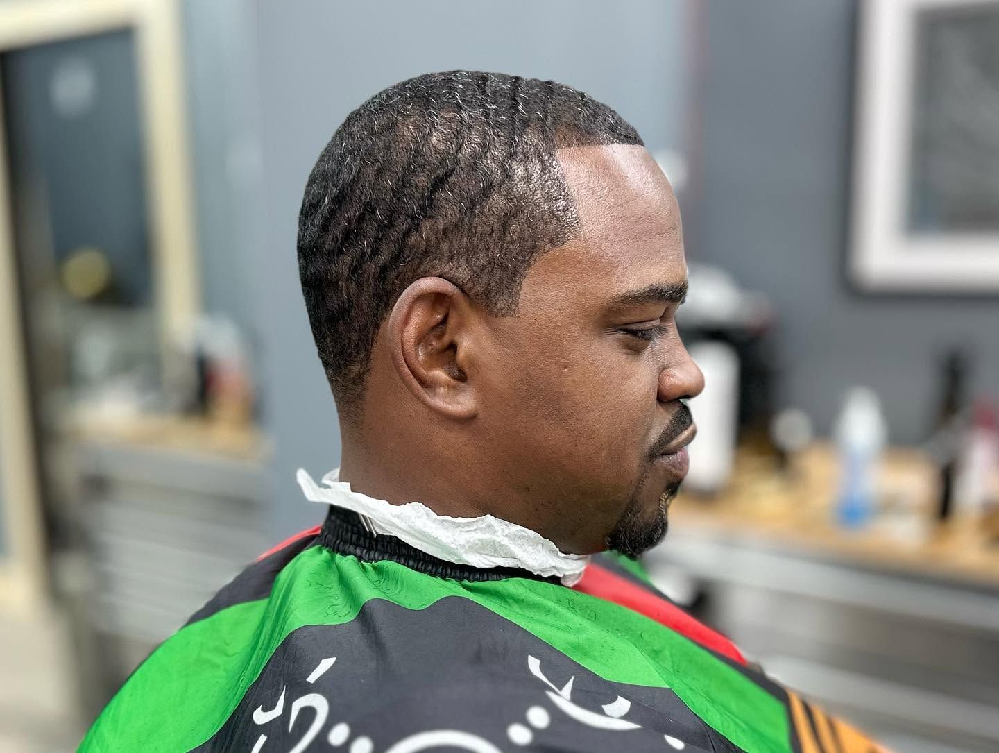 Man in a green barber cape getting a haircut in a barbershop, side profile visible