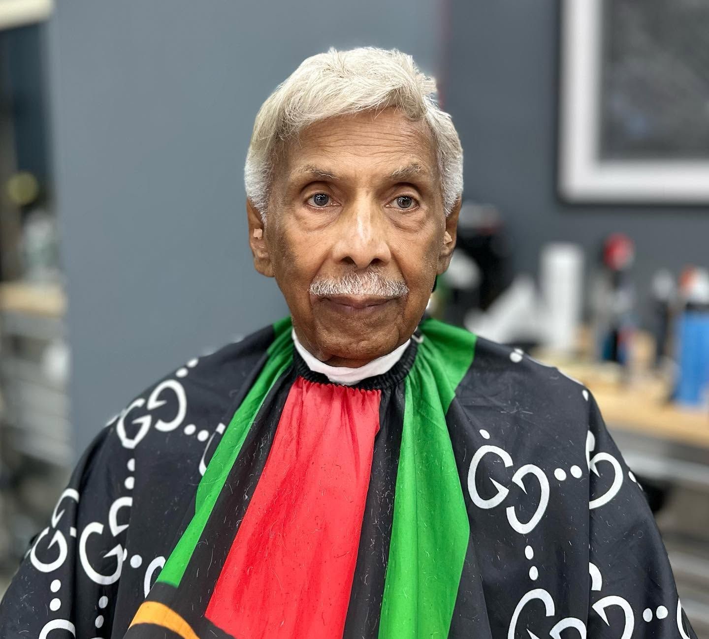 Elderly person in a black patterned shawl with green and red accents, seated indoors against a blurred background