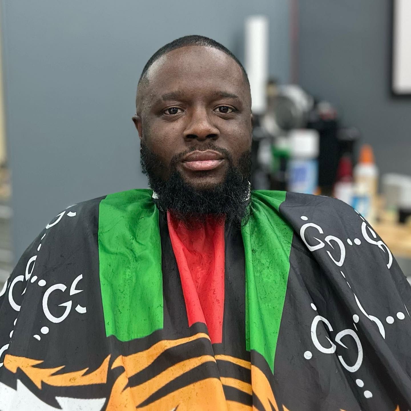 Portrait of a man in a black, green, and red barber cape, seated in a salon
