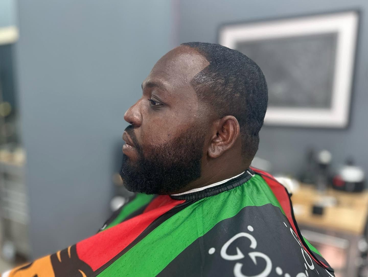 Man seated in a barber chair, wearing a red, green, and black cape, with a trimmed beard in a barbershop.