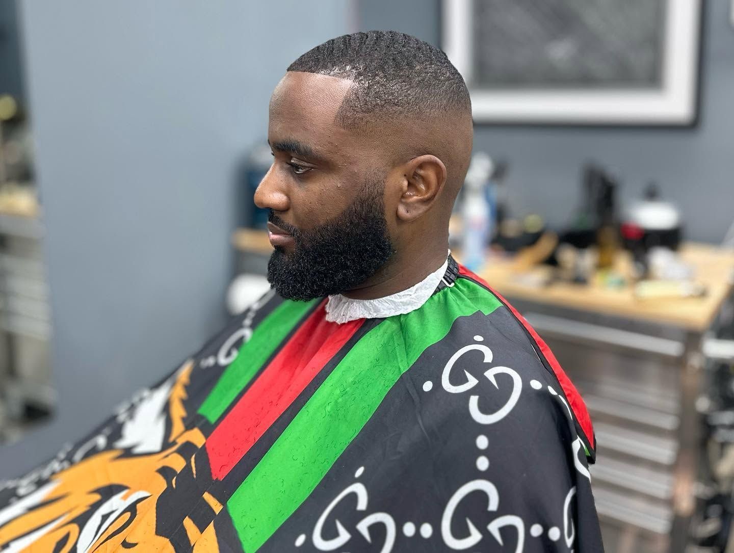 Man with a trimmed beard wearing a colorful barber cape in a salon chair, side profile, waiting for a haircut