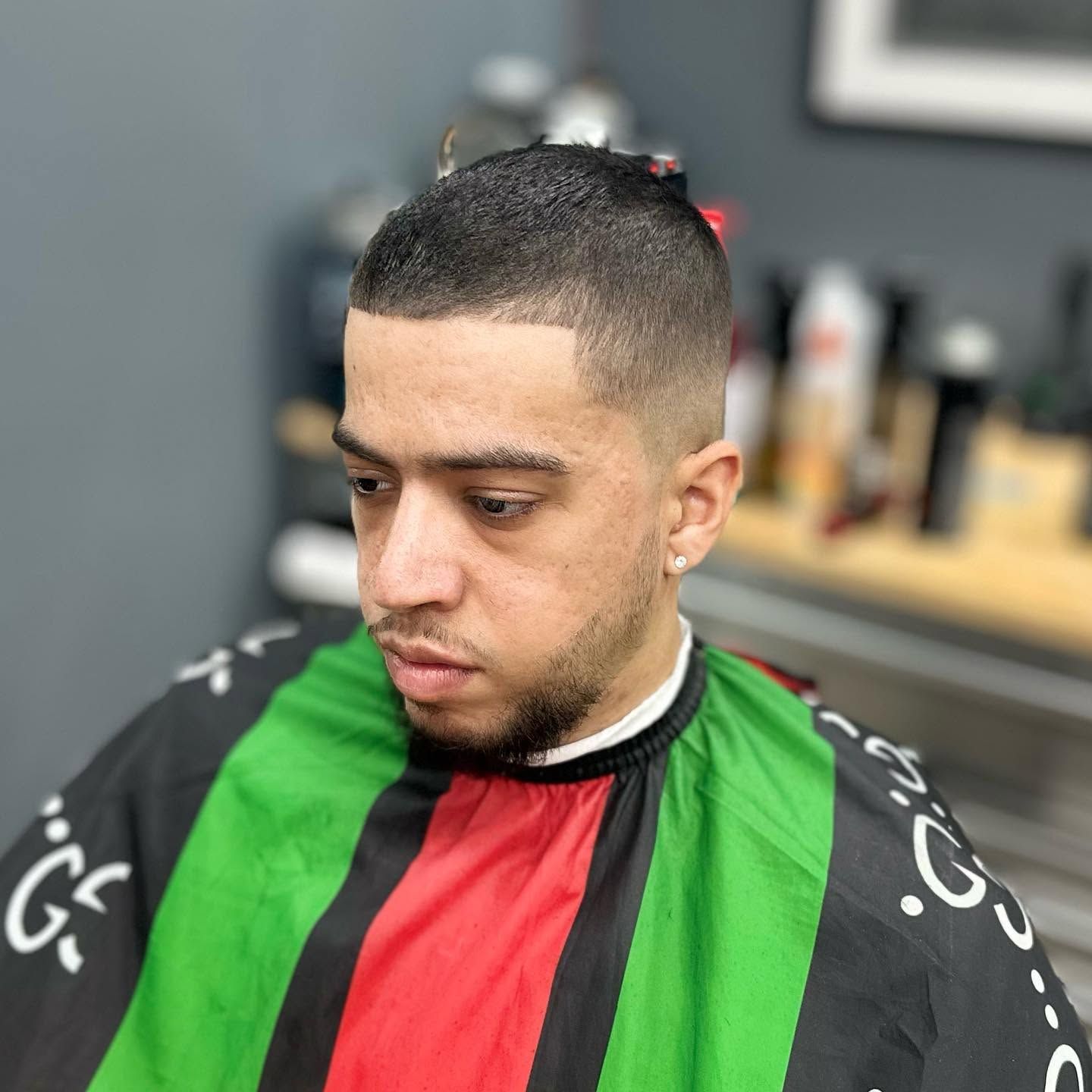 Man seated in a barber chair wearing a black, green, and red cape, with a short fade haircut.