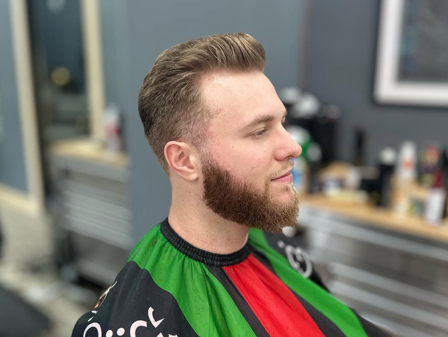Side-profile of a man with a trimmed beard in a green and red barber cape, seated in a salon chair.