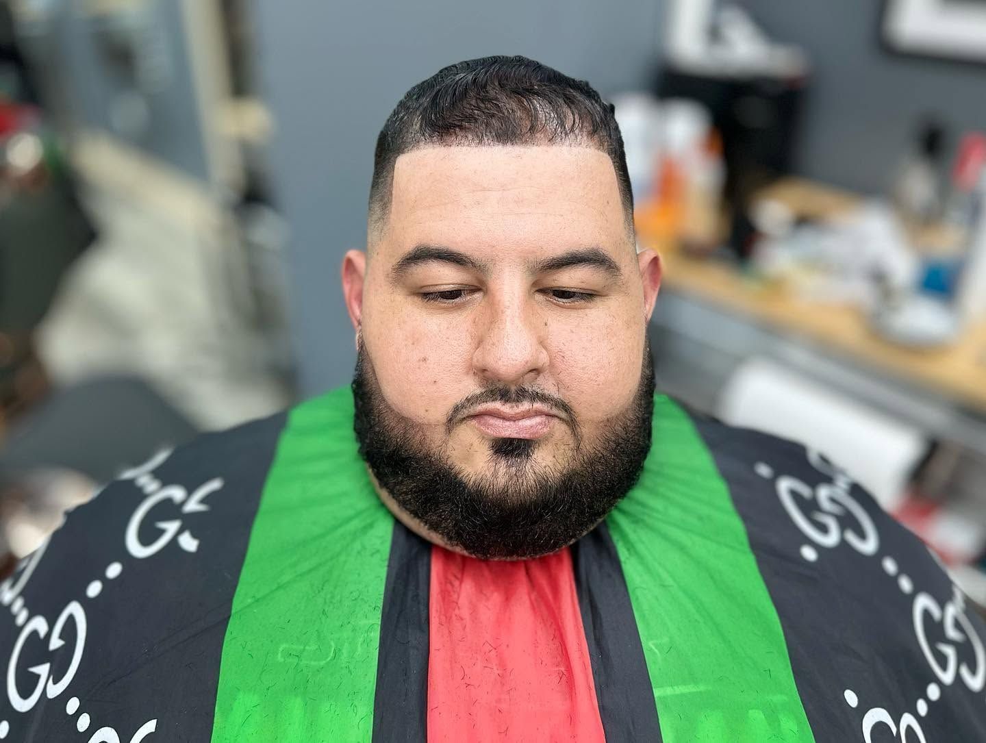 Man in black, green, and red robe with beard, looking down against a blurred indoor background