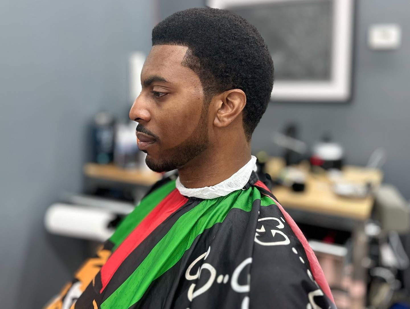Man in a barber cape with green and red stripes, shown in profile inside a barbershop.
