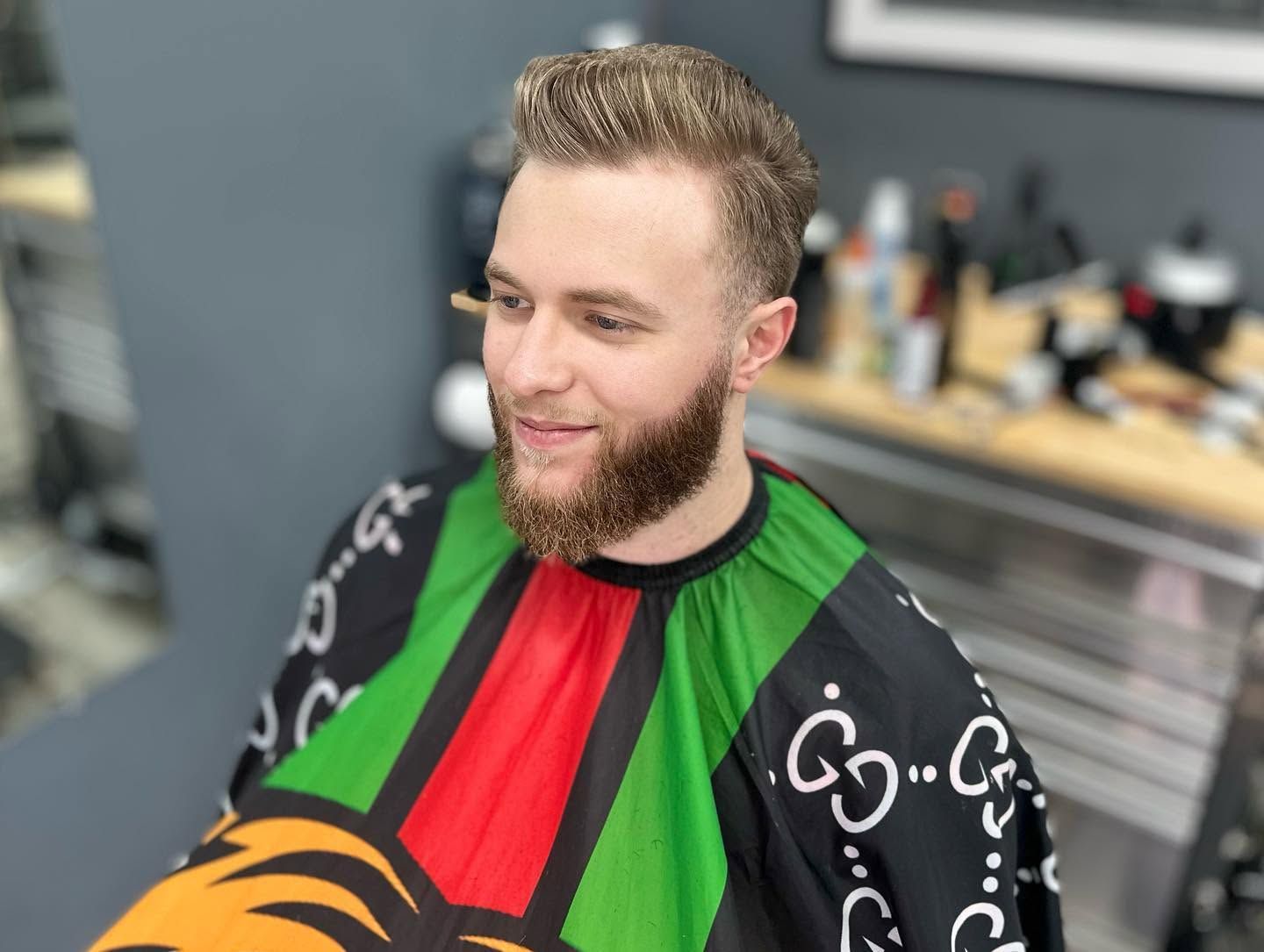 Man with beard sitting in a barber chair, wearing a colorful black, red, and green cape.
