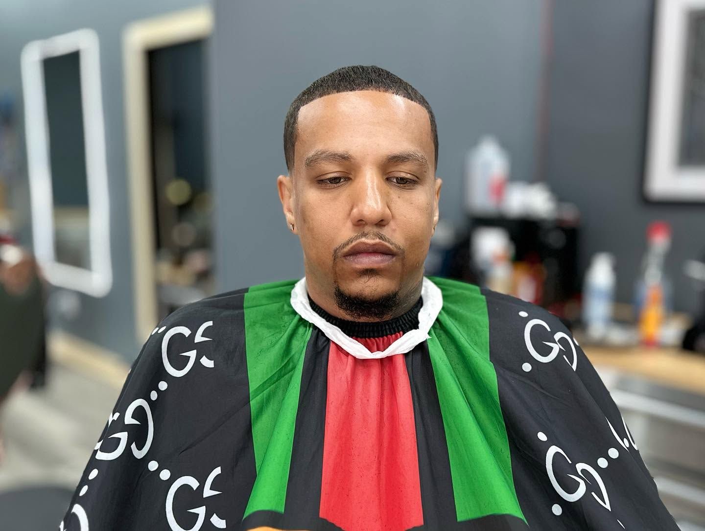 Man sitting in a barber chair wearing a black, green, and red cape, looking down with eyes closed.