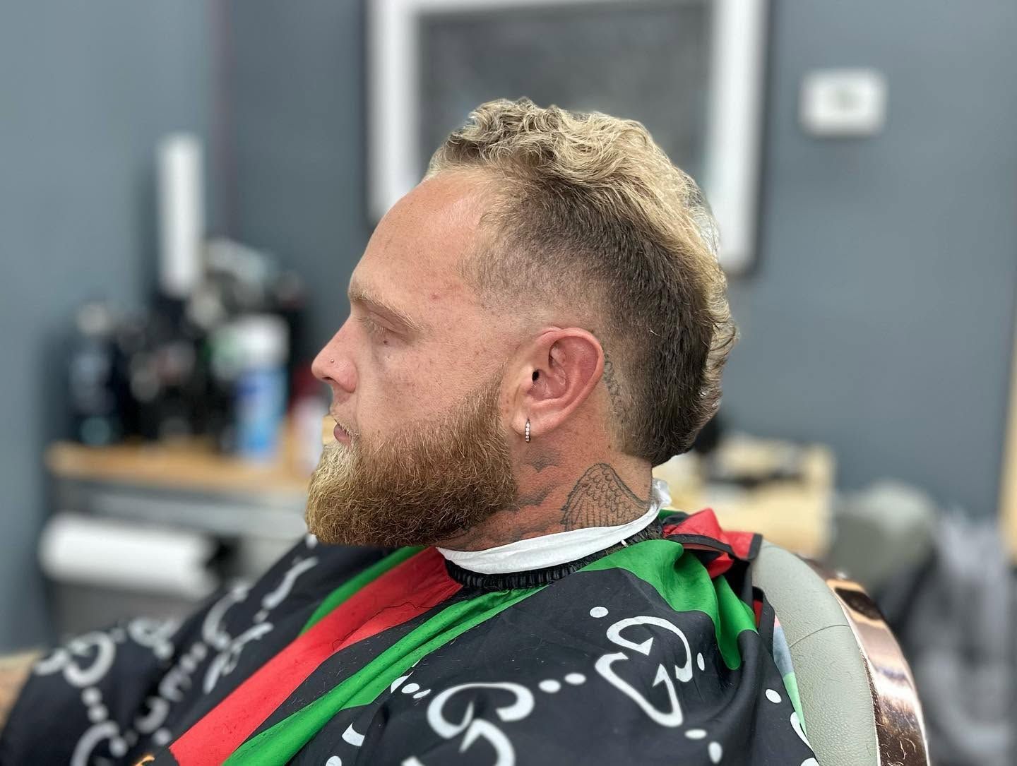 Man with a beard in a barber chair, wearing a black cape, getting a haircut in a salon