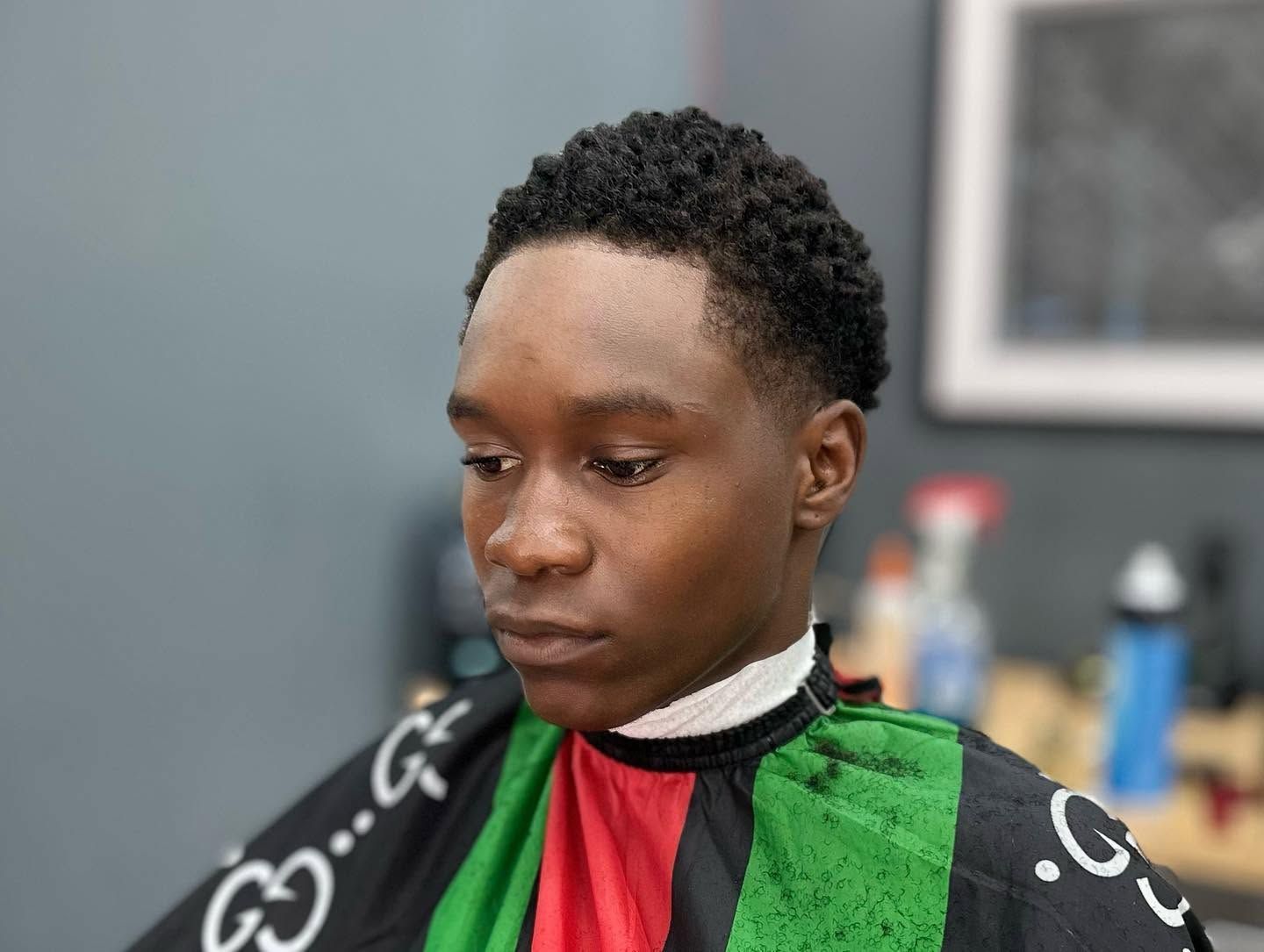 Person with short curly hair seated in a barbershop cape, looking down during a haircut