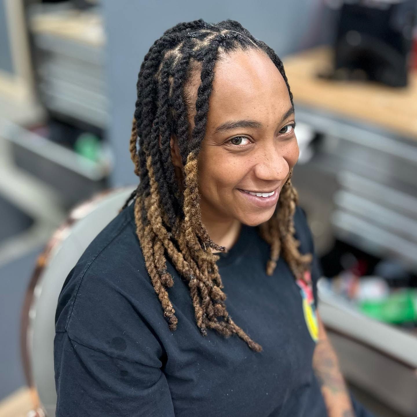 Smiling person with long braided hair in a black shirt, standing in a kitchen or workspace.
