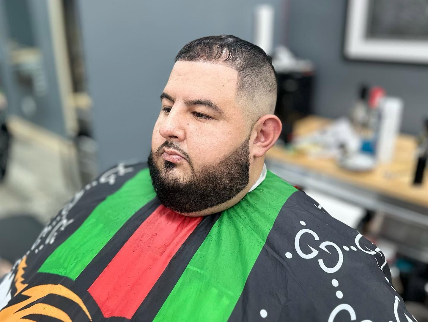 Man with a trimmed beard wearing a green, red, and black barber cape in a salon chair