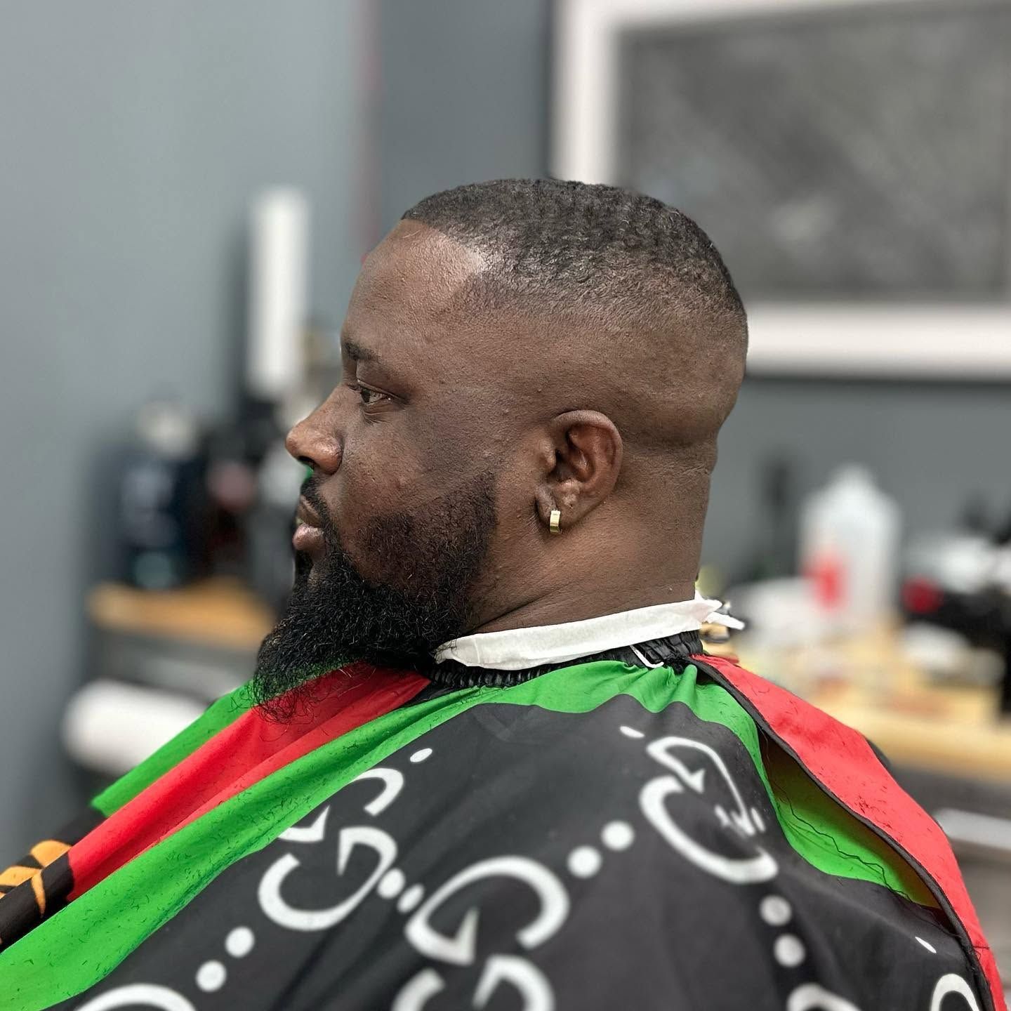 Man seated in a barber chair wearing a black cape with green and red stripes, shown in side profile.
