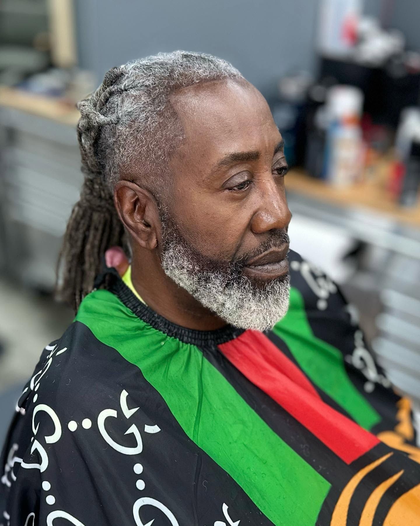 Man with gray dreadlocks and beard wearing a black, green, and red barber cape, seated in a salon chair.