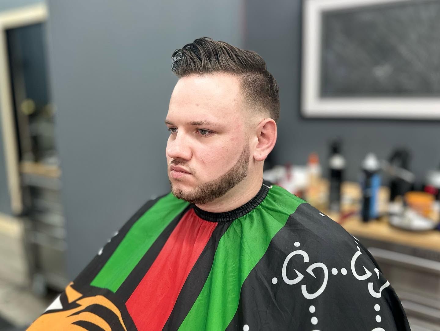 Man in a black and red barber cape with green accents, seated in a barbershop, looking left.