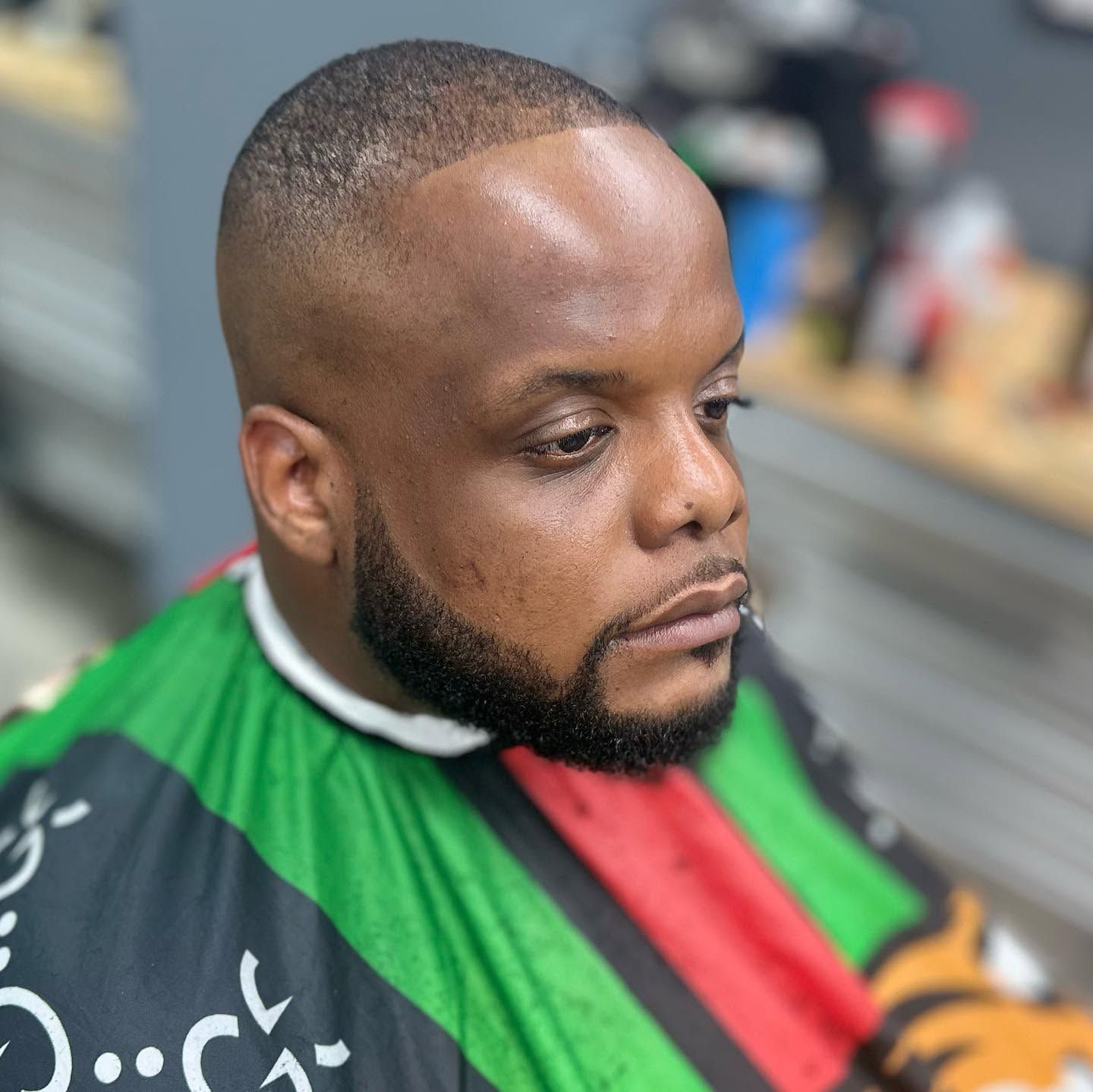 Man seated in a barber chair wearing a green, red, and black cape, with a fresh haircut and beard trim
