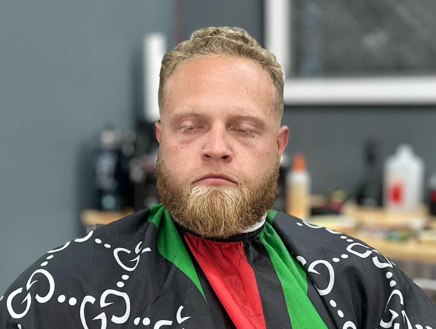 Man with blond beard in a black patterned barber cape, eyes closed, seated in a salon chair.