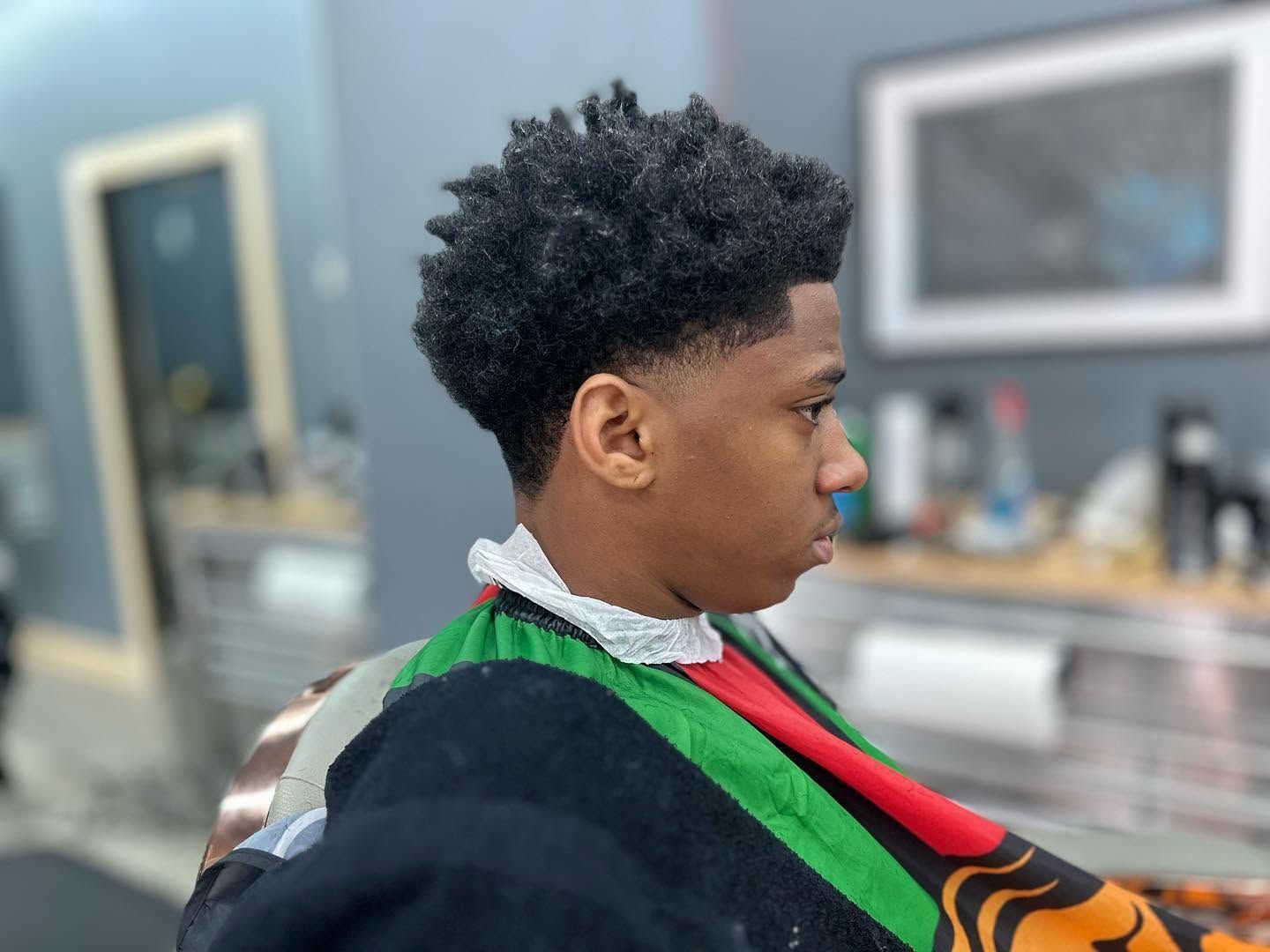Person with short curly hair in a barber chair, wearing a striped cape, side profile in a salon.