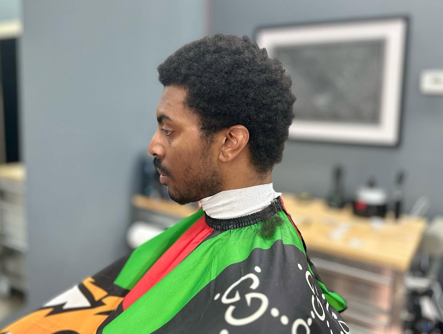 Person in a green and red barber cape, shown in profile inside a barbershop.