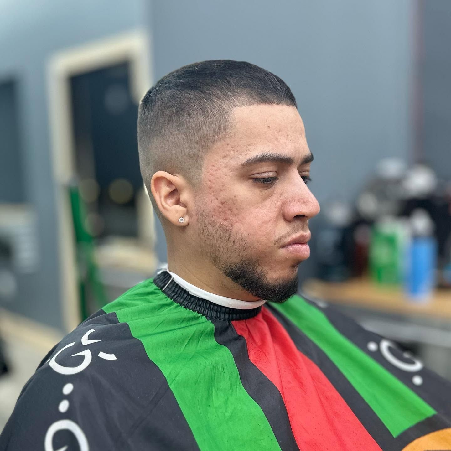 Man in a barber chair wearing a green, red, and black cape, looking down in a salon.