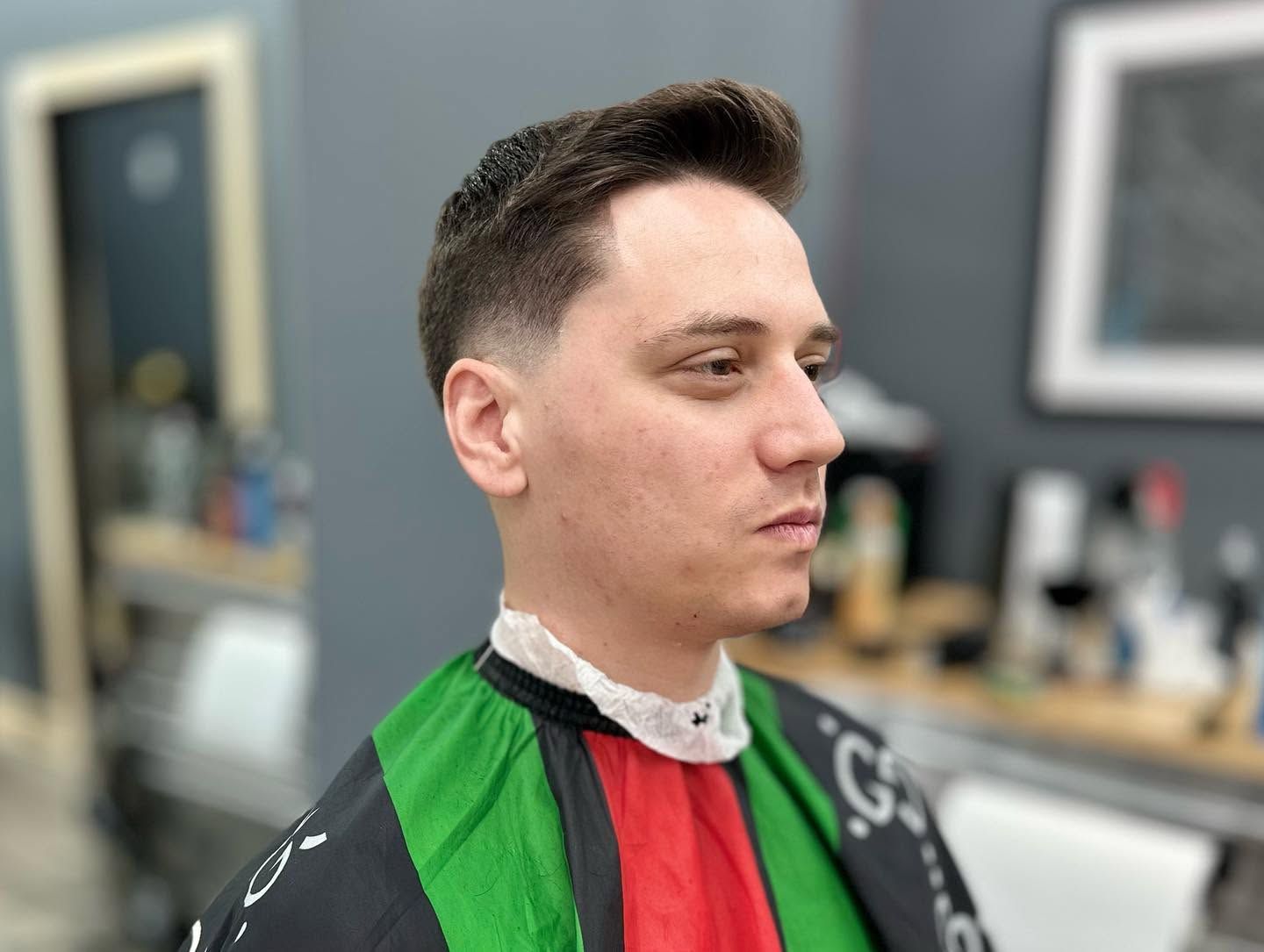 Person in a black, green, and red barber cape sitting in a salon, seen in profile.