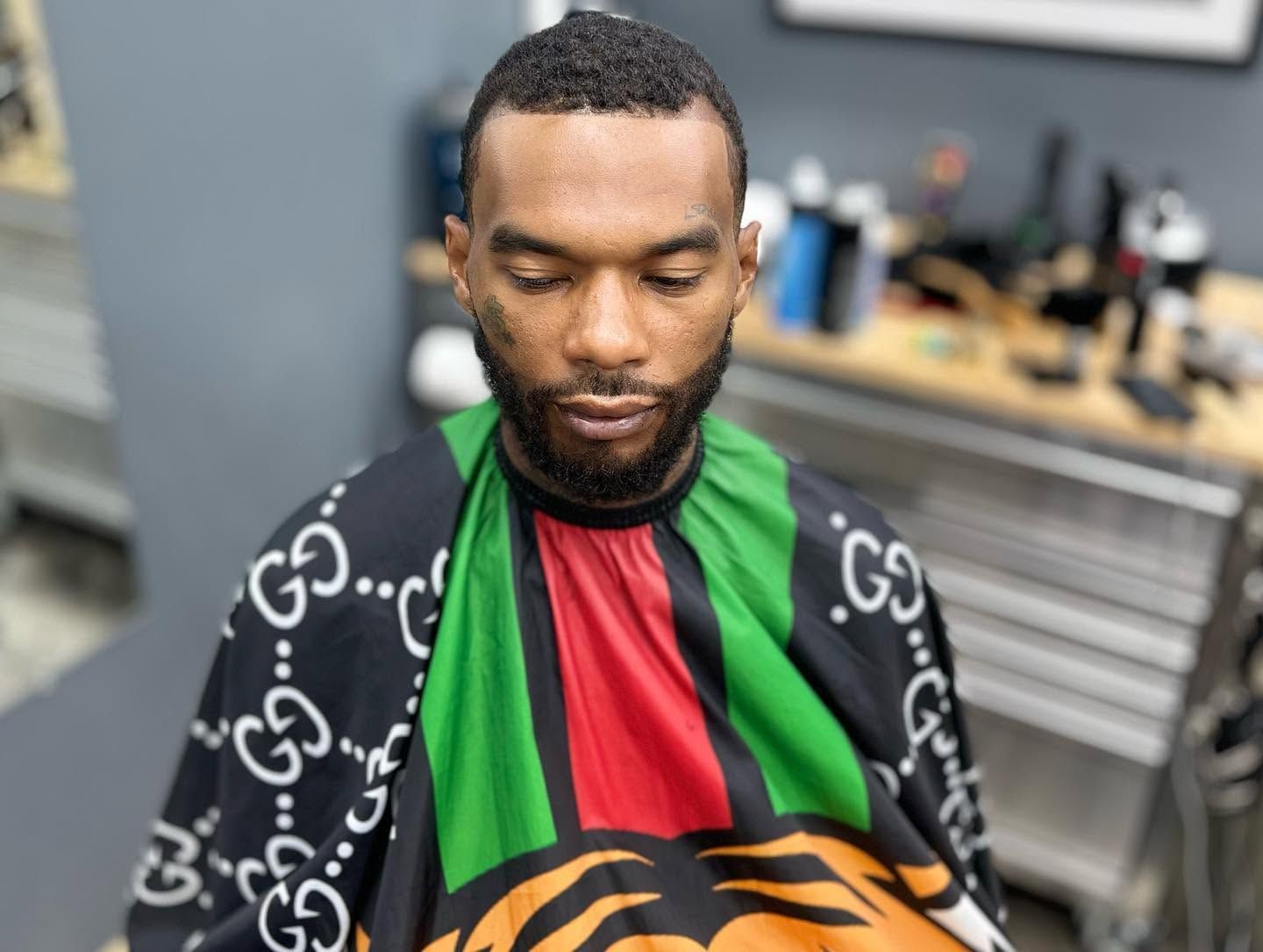Man seated in a barber chair with a black, green, red, and orange haircut cape in a barbershop