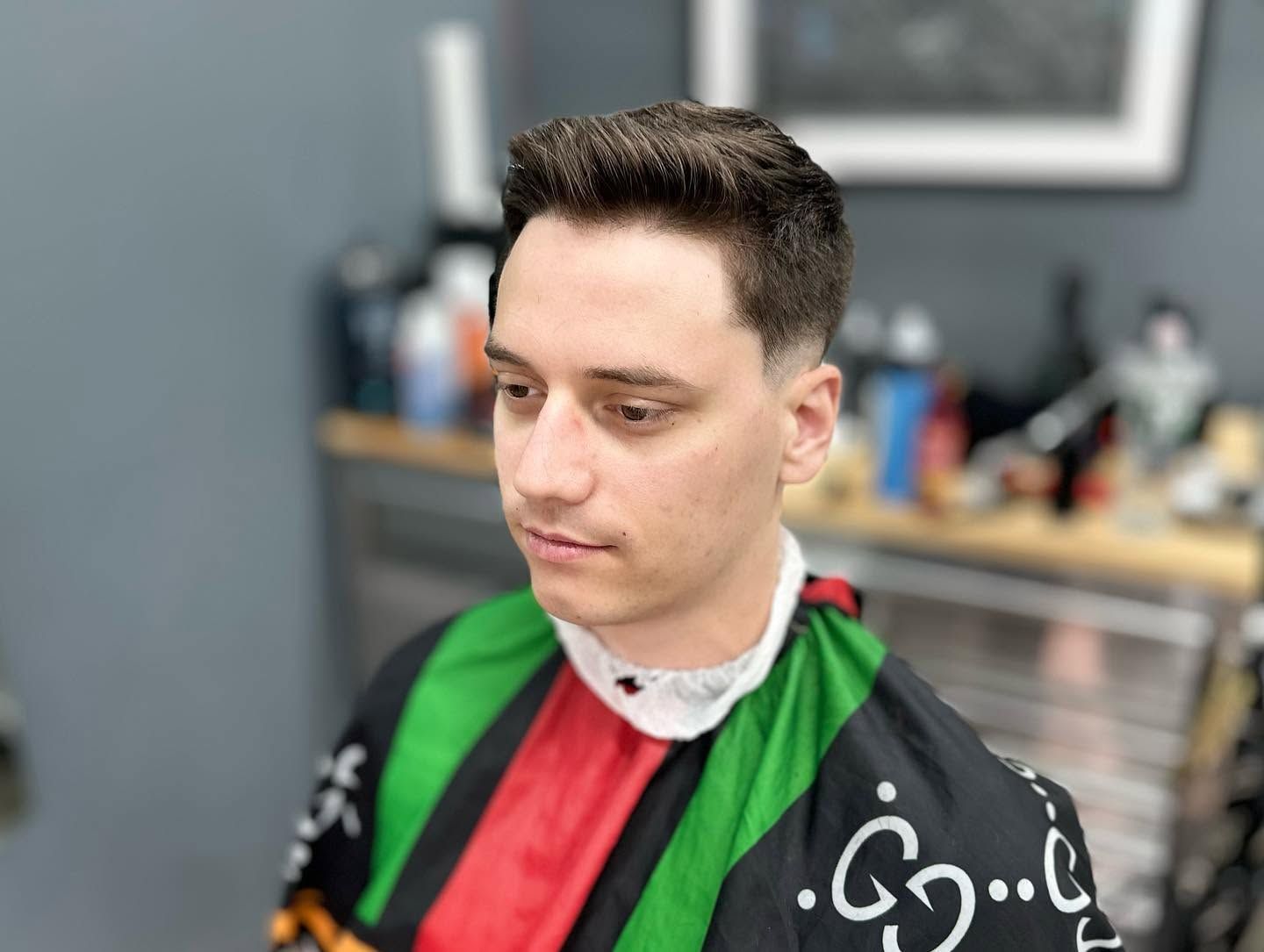Person in a barber cape with green, red, and black stripes, seated in a salon, looking down.