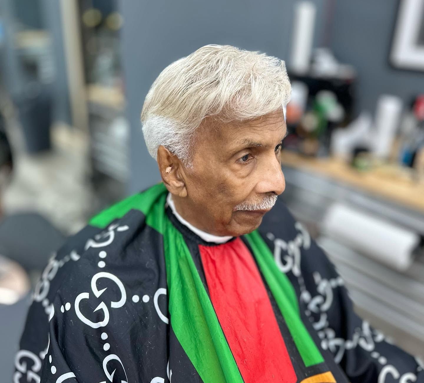 Portrait of a person with white hair wearing a black patterned coat and bright green and red scarf indoors