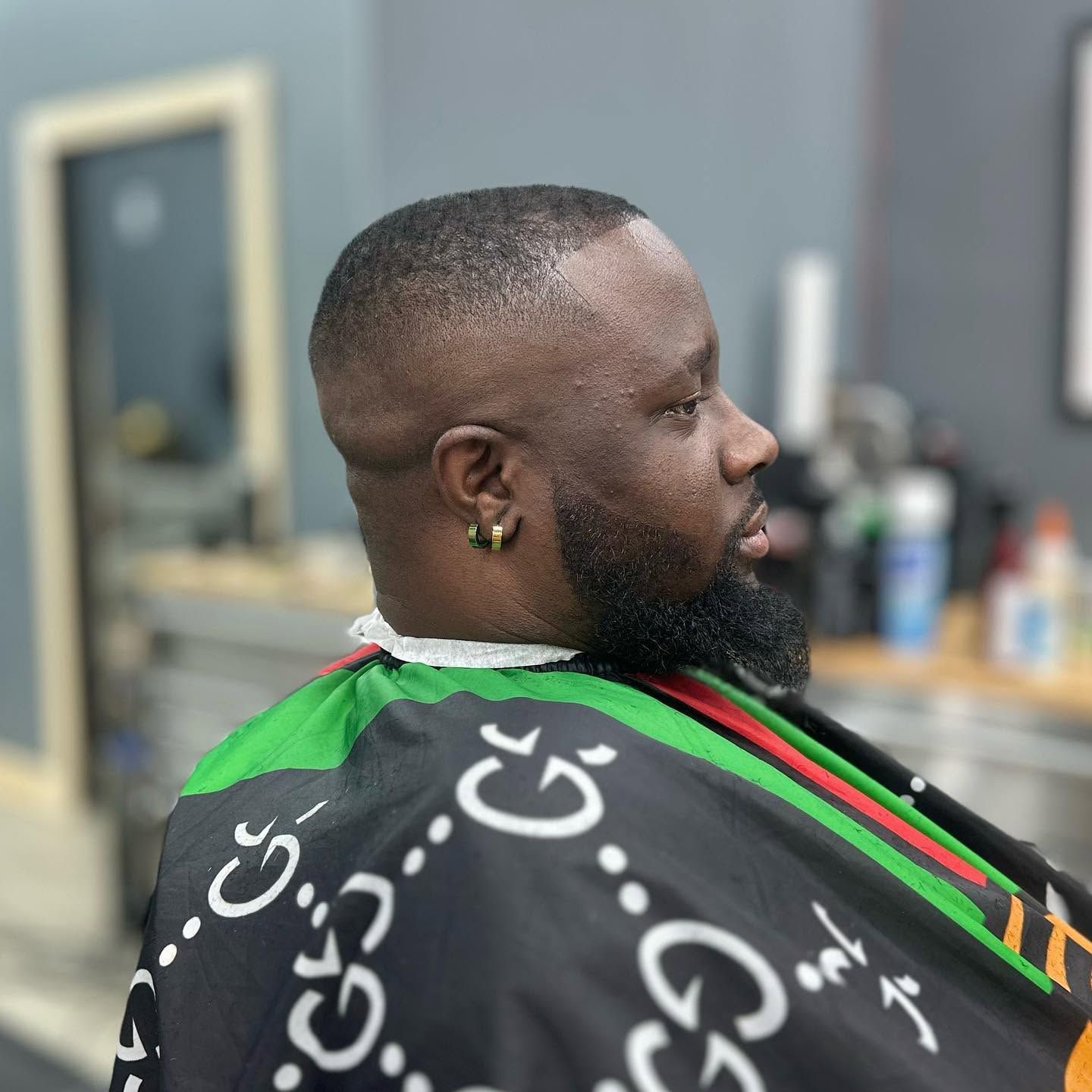 Man seated in a barbershop chair, side profile, wearing a black cape with green and red stripes.