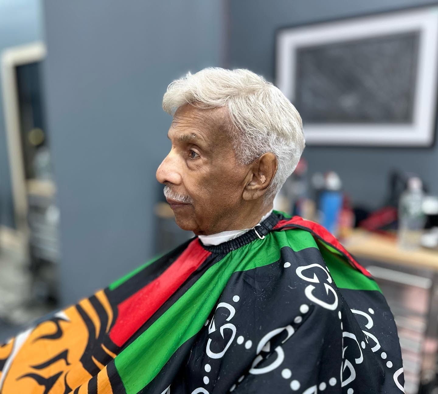 Person seated in a barbershop cape with black, green, red, and orange patterns, in profile view.