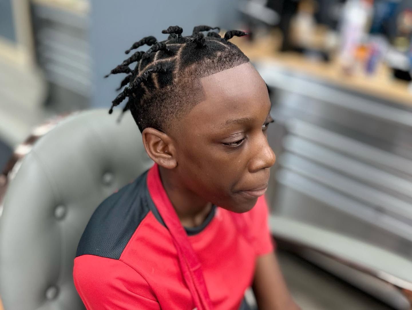Person with braided hair in a red shirt sitting in a salon chair, looking to the side.
